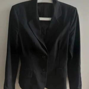 Svart kavaj från Mexx - Snygg svart kavaj från Mexx med klassisk passform och två knappar framtill. Fodret är i blankt material och kavajen har fickor fram. Diskret broderad Mexx-logga på ärmen ger en cool detalj. Perfekt för dig som gillar stilrena plagg. Har kavajen även i storlek M