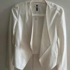 Vit kavaj från H&M med öppen front - Stilren vit kavaj från H&M med öppen front och snyggt skuren nederkant. Långärmad modell i mjukt tyg som ger en clean och fräsch look. Perfekt för dig som vill ha en enkel men trendig jacka till din outfit