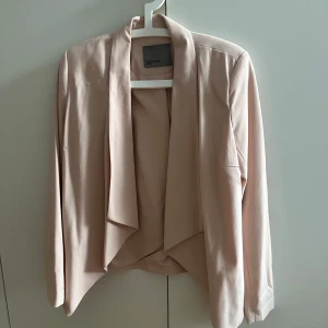 Puderrosa kavaj från Vero Moda - Snygg puderrosa kavaj från Vero Moda. Kavajen har en öppen framsida utan knappar och ett mjukt, lätt tyg som ger ett avslappnat men stilrent intryck. Perfekt för att lyfta vilken outfit som helst