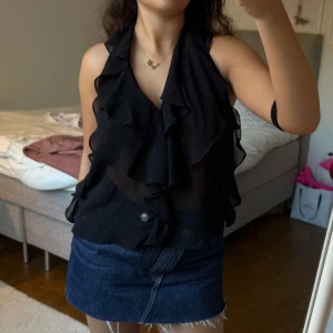 Svart volangig blus med v-ringning - Svart blus med v-ringning och volanger. Genomskinlig modell och lite öppen i ryggen. Perfekt till utekvällar