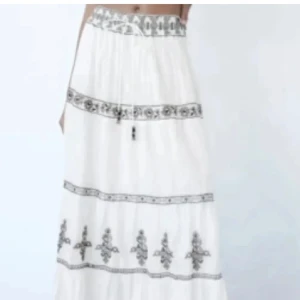 Vit långkjol med broderade detaljer - Säljer en vit långkjol med snygga grå broderade mönster och dekorativa band. Kjolen har en luftig och flowy passform, perfekt för varma dagar. Den har snörning i midjan för att justera passformen och är tillverkad i ett mjukt bomullstyg.