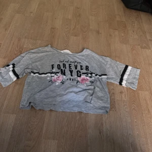 Grå croppad topp Forever NYC - Grå croppad topp med trycket 'Forever NYC 1998' och texten 'tout est magique' framtill. Toppen har blommigt mönster med rosa rosor och svarta samt vita ränder på ärmarna. Modellen är loose fit med trekvartsärm och rund hals.