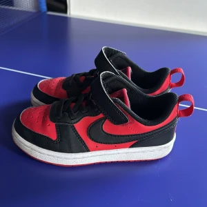 Nike sneakers röd/svart barnstorlek 28 - Snygga Nike sneakers i rött och svart med klassisk swoosh på sidan. Skorna har både kardborreband och svarta snören för enkel påtagning. Ovandelen är i syntet och textil, med vit sula och ventilerande hål på tån. Perfekta för en aktiv vardag.