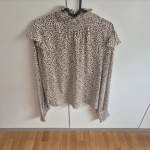 Beige blus med leopardmönster och volang - Supersnygg beige blus med svart leopardmönster, hög smockad krage och långa ärmar med breda smockade muddar. Blusen har volangdetaljer vid axlarna och knäppning bak i nacken. Perfekt för dig som gillar trendiga och unika plagg.