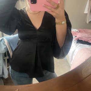 Svart blus med vida ärmar och v-ringning - Säljer en svart blus med snygg v-ringning och riktigt vida ärmar som ger en cool siluett. Blusen har en lätt glansig finish och ett veck framtill som ger extra form. Perfekt att styla med jeans eller kjol för en trendig look.