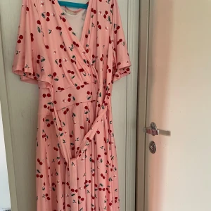 Rosa omlottklänning med körsbär - Söt rosa omlottklänning med körsbärsmönster och korta ärmar. Klänningen har v-ringad hals och ett knytband i midjan som ger en snygg siluett. Perfekt för dig som gillar färgglada och lekfulla plagg med retrovibbar. Kollektionen är Ellos x Linda-Marie