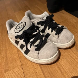 Adidas Campus sneakers i grå mocka - Snygga Adidas Campus sneakers i ljusgrå mocka med svarta ränder och svart snörning. Klassisk rund tå och platt sula med gummisula i brunt. Stor Adidas-logga där fram och Campus-tryck på sidan. Säljer pga använder ej, de har ej defekter 