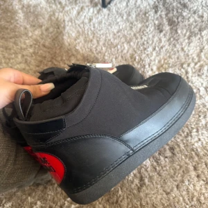 Love Moschino svarta boots med hjärta - Svarta boots från Love Moschino med chunky platt sula och röda hjärtformade detaljer bak. Skorna har dragkedja framtill med vit och röd hjärtdetalj samt textlogga. Materialmix av skinn och syntet, vadderad insida för extra komfort. ÄKTA MOSCHINO,  nypris 2000 kr, säljs för 1100 men man kan diskutera ALDRIG använt 