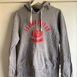 Grå hoodie - Snygg grå hoodie med huva och vita snören. Framtill står det 'Leisurewear 87' i rött med broderade detaljer. Tröjan har känguruficka och ribbade muddar. Perfekt för en avslappnad och sportig look.