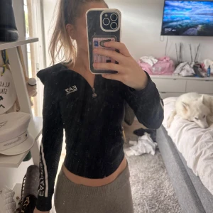 Svart EA7 hoodie med dragkedja - Svart croppad hoodie från EA7 Emporio Armani med vit logga på bröstet och längs ärmarna. Tröjan har dragkedja framtill, huva och långa ärmar. Snygg och sportig look med vita detaljer som ger extra edge. Den är omsydd för att sitta extra tight i midjan! Köpt för 1700kr