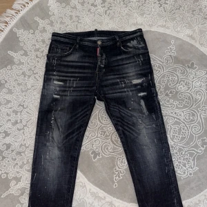Svarta jeansbyxor från Dsquared2 ICON - Säljer ett par svarta Dsquared2 ICON jeans med slitningar och färgstänk. Byxorna har fem fickor, knappgylf och en tydlig Dsquared2-logga bak i midjan. (Ni som kan märken ser att detta är en äkta vara)