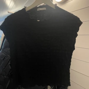 Svart strukturerad topp från Zara - Svart topp från Zara med strukturerad, lätt glansig yta och vågiga kanter vid hals och ärm. Passar som en S! Modellen är ärmlös och har en croppad passform som ger en trendig vibe. Perfekt att styla med jeans eller kjol för en cool look.