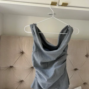 Grå ärmlös draperad topp - Snygg grå topp med draperad front och breda axelband. Toppen är ärmlös och har en cool, lite asymmetrisk look som passar perfekt till jeans eller kjol. Materialet känns mjukt och har en lätt struktur.
