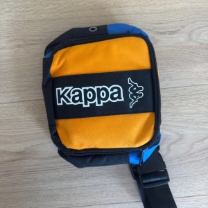 Kappa axelväska i gult, blått och svart - Säljer en färgstark axelväska från Kappa med cool logga framtill. Väskan har flera fack med dragkedja, justerbar rem och är gjord i slitstark polyester. Perfekt för att bära dina viktigaste grejer och ge outfiten en sportig vibe.
