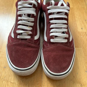 Vinröda Vans Old Skool sneakers - Klassiska Vans Old Skool sneakers i vinrött med vit sula och vit sidosnirk. Ovandel i mocka och canvas, vita snören och gummisula med det ikoniska rutmönstret undertill. Rund tå och låg modell, perfekta för en avslappnad streetstil.