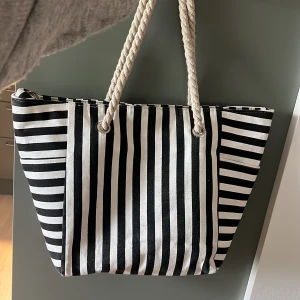 Randig strandväska med rep-handtag - Stor svartvit shoppingväska med breda och smala ränder. Väskan har rejäla rep-handtag i vitt och är tillverkad i ett kraftigt bomullstyg. Perfekt för att bära med sig allt man behöver till stranden eller stan.