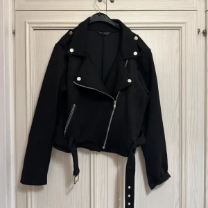 Svart bikerjacka med bälte och dragkedja - Snygg svart bikerjacka med silvriga dragkedjor, stora slag och bälte i midjan. Jackan har långa ärmar och coola detaljer som knappar på axlarna. Perfekt för dig som vill ha en edgy stil.