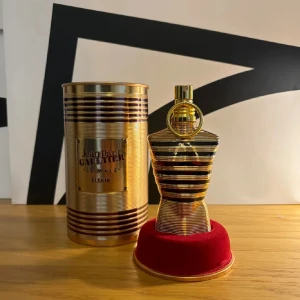 Jean Paul Gaultier Le Male Elixir - Exklusiv parfymflaska i guld och svart från Jean Paul Gaultier, Le Male Elixir. Flaskan har en ikonisk bystform med horisontella ränder och levereras med en lyxig guldfärgad metallburk och röd sammetsbas. Perfekt för dig som gillar unika och lyxiga dofter.