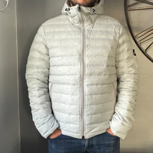 Ljusgrå dunjacka från J.Lindeberg - Snygg ljusgrå dunjacka från J.Lindeberg med huva och dragkedja framtill. Jackan har quiltad design, två fickor med dragkedja och logga på ärmen. Perfekt för kyliga dagar när du vill hålla stilen och värmen.