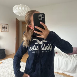 Marinblå Hollister hoodie med foder - Mysig marinblå hoodie från Hollister med vit broderad logga och text på bröstet. Tröjan har dragkedja, snörning i huvan och fluffigt vitt foder på insidan av huvan. Perfekt för kyliga dagar och har två fickor framtill😊
