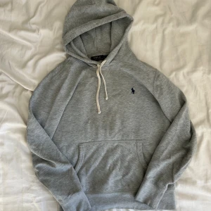 Grå hoodie från Polo Ralph Lauren - Klassisk grå hoodie från Polo Ralph Lauren med broderad marinblå logga på bröstet. Tröjan har huva med vit snörning, känguruficka och långa ärmar. Perfekt för en chill och stilren look. Tillverkad i mjuk bomullsmix för extra komfort.