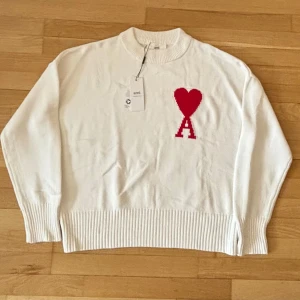 Vit Ami Paris Tröja - Vit stickad tröja från Ami Paris med röd hjärtlogga och bokstaven A på bröstet. Tröjan har rund hals, ribbade muddar och nederkant samt lång ärm. Perfekt för dig som gillar stilrena plagg med en twist. Tveka inte för att ställa frågor.