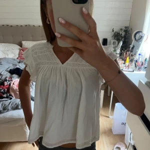 Vit blus med volang från Gina Tricot - Supersöt vit blus från Gina Tricot med v-ringning, volangdetaljer på ärmarna och fina broderade partier upptill. Luftig och lätt i bomull, perfekt för varma dagar. Passar dig som gillar romantisk och avslappnad stil.