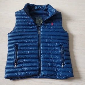 mörk Blå dunväst från Polo Ralph Lauren - Snygg marinblå dunväst från Polo Ralph Lauren med horisontella quiltade sömmar och dragkedja framtill. Västen har två fickor med dragkedja och klassisk röd broderad logga på bröstet. Perfekt att slänga över en hoodie för en chill look.OBS! Har tyvärr tappat bort lappen.