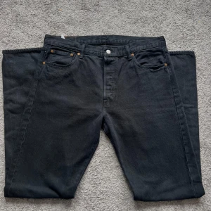 Svarta Levi's 501 jeans W36 L36 - Klassiska svarta Levi's 501 jeans med rak passform och fem fickor. Jeansen har knappgylf, ikonisk läderpatch bak och är tillverkade i robust bomullsdenim. Perfekta för dig som gillar tidlös stil och vill ha ett par jeans som funkar till allt.