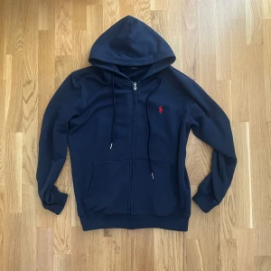 Marinblå hoodie från Polo Ralph Lauren - Säljer en marinblå hoodie från Polo Ralph Lauren med dragkedja och huva. Tröjan har en röd broderad logga på bröstet, känguruficka och snörning i huvan. Perfekt för chill dagar och snygg till jeans eller joggers.