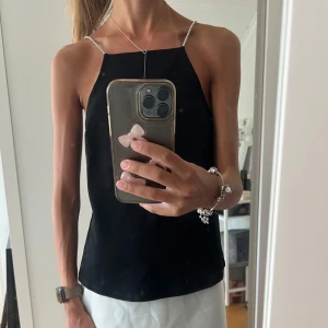 Svart topp med glittriga axelband  - Stilren svart linnetopp från H&M med tunna, glittriga axelband. Toppen har rak halsringning och är tillverkad i 95% återvunnen polyester. Perfekt för dig som gillar minimalistisk och trendig stil. Aldrig använd prislappen sitter kvar. 