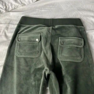 Gröna mjukisbyxor i velour JC - Mysiga gröna mjukisbyxor i velour från Juicy Couture. Byxorna har bred resår i midjan med snörning, två stora fickor med lock och knappar bak samt broderad JC-logga framtill. Perfekta för chill eller streetwear.