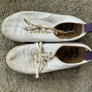 Vita Eytys Fugu sneakers - Snygga Eytys Fugu sneakers i vitt canvasmaterial med klassisk rund tå och platt sula. Skorna har guldfärgade öljetter, vita snören och en lila hälflik. Perfekta för dig som gillar minimalistisk och stilren design.