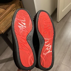 Christian Louboutin svarta sneakers med nitar - Säljer ett par svarta sneakers från Christian Louboutin med ikonisk röd sula och svarta nitar över tån. Skorna har snörning och är tillverkade i skinn med detaljer i mocka. Insidan är ljusbeige och sulan har Louboutin-loggan.