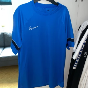 Blå Nike fotbollströja Dri-FIT M - Snygg blå fotbollströja från Nike i Dri-FIT-material som andas. Klassisk design med korta ärmar, svart detalj på ärmarna och vit Nike-logga på bröstet. Perfekt för träning eller match. Skön passform och sportig look.