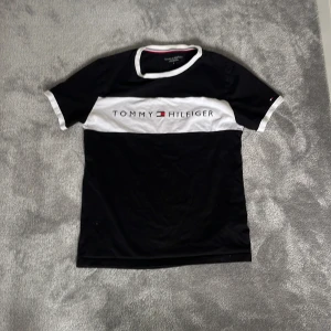 Svart och vit t-shirt Tommy Hilfiger - Svart t-shirt från Tommy Hilfiger med logga över bröstet. Klassisk rund halsringning och vita detaljer vid ärmslut och krage👕 Skön passform och stilren design, perfekt till jeans eller shorts på sommaren☀️