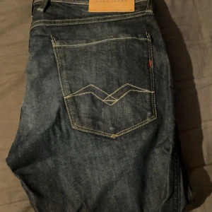 Replay jeans i mörkblå denim - Säljer ett par snygga Replay jeans i mörkblå denim med klassiska bakfickor och kontrastsömmar. Jeansen har rak passform och normal midja, perfekt för dig som gillar stilrena och tidlösa jeans. Märkespatch i läder baktill.