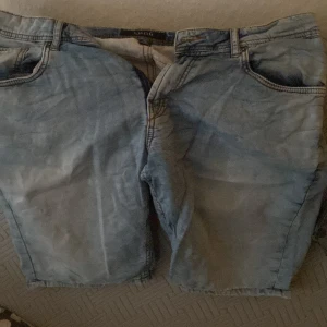 Ljusblå jeansshorts från SMOG - Ljusblå jeansshorts från SMOG med klassisk femficksdesign och råa benslut. Shortsen har normal passform och bälteshällor i midjan. Perfekta för varma dagar och en avslappnad stil.
