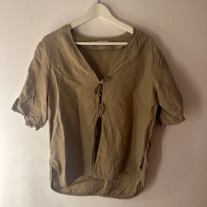 Sagegrön blus med knyt från Samsøe Samsøe - Snygg sagegrön blus från Samsøe Samsøe med v-ringning och två knyt framtill. Modellen har korta ärmar med resår och är tillverkad i ett lätt bomullsmaterial. Perfekt för dig som gillar en avslappnad men trendig look.