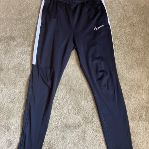 Svarta Nike träningsbyxor med vita detaljer - Svarta träningsbyxor från Nike med vita ränder längs sidorna och klassisk Nike-logga på låret. Byxorna har elastisk midja och smal passform, perfekta för sport eller chill. Lätta och bekväma i syntetmaterial.