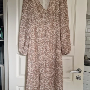 Beige blommig långklänning BikBok XS - Långklänning från BikBok i beige med vitt blommigt mönster. Klänningen har v-ringning, långa ärmar med resår vid handlederna och en slits framtill. Tillverkad i återvunnet material, luftig och perfekt för varmare dagar. Så fin på bara använda vid ett tillfälle.
