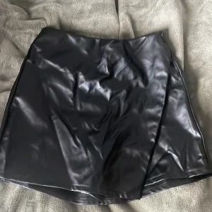 Svart kjol från Zara med shorts bakom  - Snygg svart kortkjol från Zara med shorts bakom i skinnimitation med elastisk midja. Kjolen har en stilren look och är perfekt för dig som vill ha en edgy vibe. Passar till många olika outfits och är lätt att matcha med både sneakers och boots.
