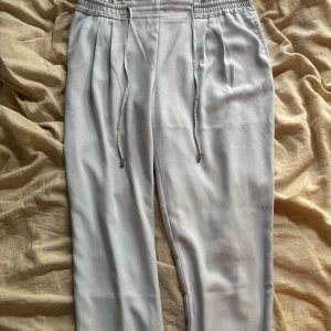 Ljusgrå byxor med snörning från Zara - Snygga ljusgrå byxor från Zara Basic med elastisk midja och snörning. Byxorna har raka ben, diskreta fickor bak och är gjorda i ett mjukt, tunt material som känns skönt mot huden. Perfekta för en chill och stilren look.