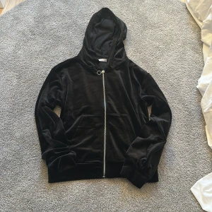 Svart sammetshoodie med dragkedja - Mjuk och stilren svart hoodie i sammet med hel dragkedja framtill och huva. Tröjan har långa ärmar och en stor ficka på magen. Perfekt för dig som gillar en chill och trendig look.