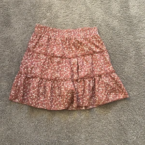 Rosa blommig volangkjol - Supersöt kort kjol i rosa med små vita och svarta blommor. Kjolen har flera volanger och resår i midjan för en bekväm passform. Perfekt för dig som gillar en romantisk och lekfull stil.