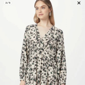 Blommig långklänning med v-ringning - Långklänning i ljusbeige med svart och grönt blommönster. Klänningen har v-ringning, långa ärmar med resår vid handlederna och en omlottliknande överdel. Materialet känns lätt och flowy, perfekt för vår och sommar.