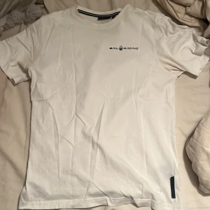 Vit t-shirt från Sail Racing - Vit t-shirt från Sail Racing med svart logga tryckt på bröstet och en liten logga i nacken. Klassisk rund halsringning och korta ärmar. Tillverkad i mjuk bomull, perfekt för en clean och stilren look. Passar om man är mellan 160-170