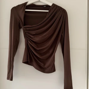 Brun topp  - Säljer en brun topp från SHEIN i storlek XS. Toppen har långa ärmar, draperad framsida och asymmetrisk skärning. Tillverkad i ett mjukt, stretchigt syntetmaterial som sitter snyggt på kroppen. Perfekt för dig som gillar trendiga detaljer och en cool look.