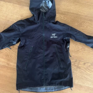 Svart vindjacka från Arc'teryx  - Svart vindjacka från Arc'teryx med huva och dragkedja framtill. Jackan har två snedställda fickor med dragkedja och diskret logga på bröstet. Tillverkad i lätt syntetmaterial, perfekt för blåsiga dagar. Snygg och funktionell design. Size Small. Köpt begagnad så vet inte om den är äkta eller ej.