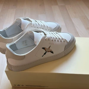 Vita sneakers från Axel Arigato - Snygga vita sneakers från Axel Arigato med broderad fågel på sidan. Skorna har klassisk låg profil, rund tå och vita snören. Tillverkade i skinn med detaljer i ljusgrå mocka vid tån. Perfekta för en clean och stilren look. Skorna är använda endast 1 gång. Klistermärken tillkommer. 